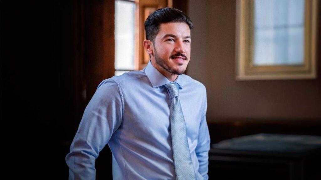 Triangulaciones-de-samuel-garcia-en-nl - Regios Al Cuadrado Smiling Man in a Light Blue Dress Shirt and Pale Tie Leans Against a Wall by a Window in a Softly Lit Room. - Regios Al Cuadrado