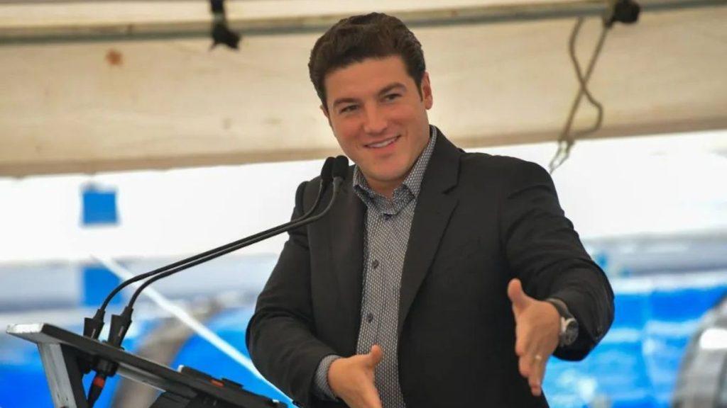 Man in a Dark Blazer Delivering a Speech at a Lectern with Two Microphones, Gesturing with Open Hands and Smiling. - Regios Al Cuadrado