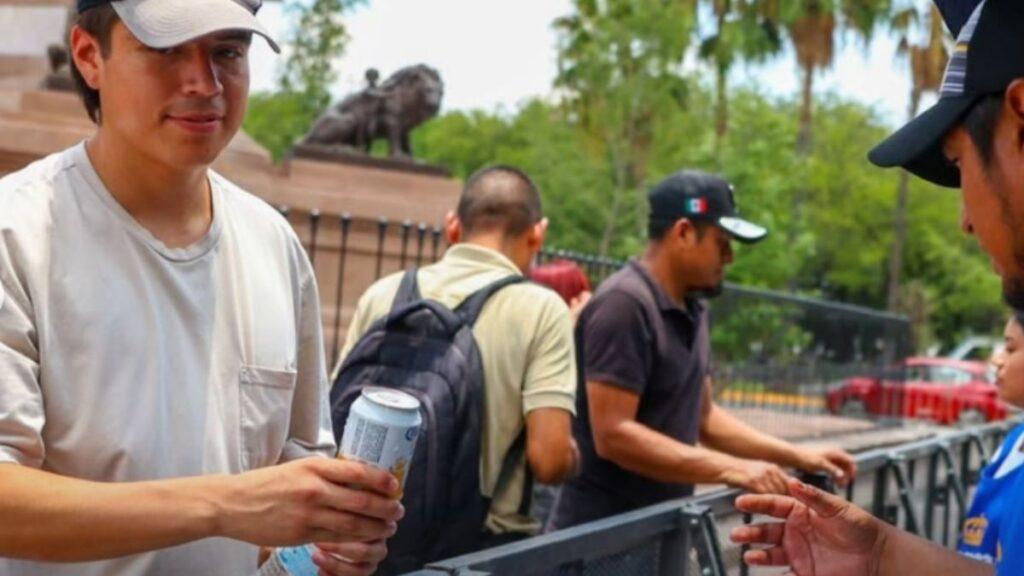 Cuanto-duro-la-entrega-de-cerveza-gratis-en-la-macroplaza - Regios Al Cuadrado Man in a Cap Hands a Can to Someone at a Railing, with Others Chatting in the Background at a Park. - Regios Al Cuadrado