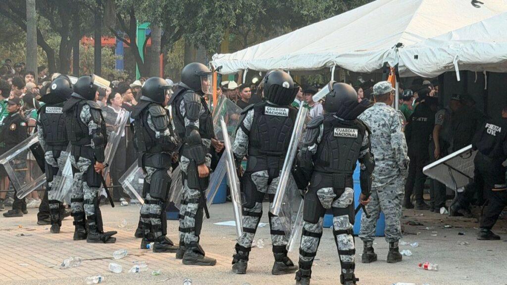 Line of Riot Police in Black Armor with Clear Shields Outside a White Tent, Facing a Crowd in a Street Scene. - Regios Al Cuadrado
