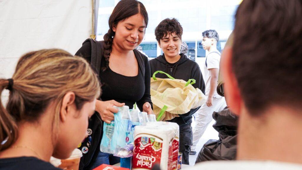 Bloque 2- Junio 32 - Regios Al Cuadrado Group of Young Volunteers Sorting and Packing Donations at a Table with Water Bottles and Paper Towels Visible in a Tent Area. - Regios Al Cuadrado