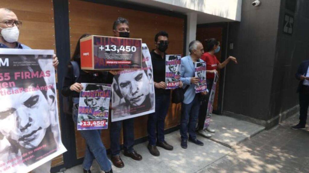 Group of Protesters Standing Outside a Building, Holding Posters with Faces and the Number 13,450; Several Wear Masks. - Regios Al Cuadrado