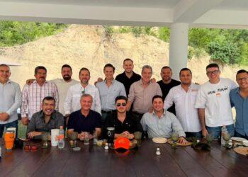 Group of Men Posing for a Photo at an Outdoor Gathering Under a Pavilion, Seated and Standing Around a Long Table with Drinks and Plates. - Regios Al Cuadrado