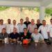 Group of Men Posing for a Photo at an Outdoor Gathering Under a Pavilion, Seated and Standing Around a Long Table with Drinks and Plates. - Regios Al Cuadrado