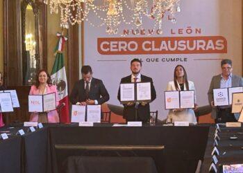 Group of Officials in a Formal Meeting Holding Signed Documents, Standing Behind a Long Table in Front of a Banner That Reads 'cero Clausuras' with a Mexican Flag Nearby. - Regios Al Cuadrado